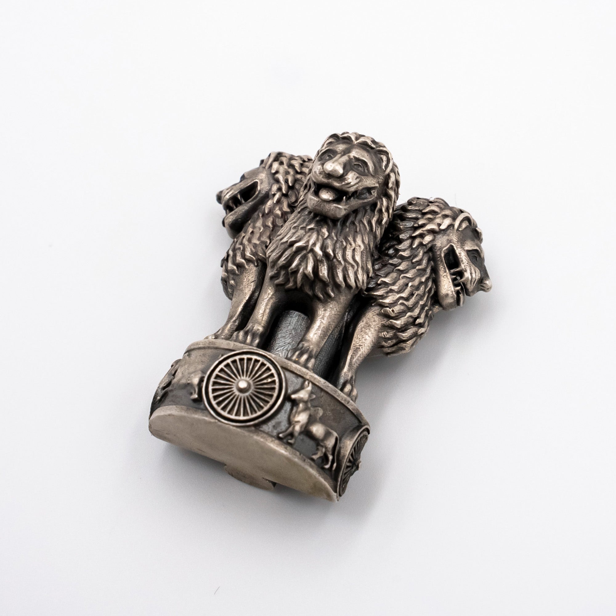 A metallic silver-toned sculpture of the Ashoka Chakra, featuring three lions standing on a wheel, photographed on a clean white background.